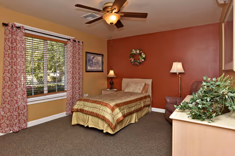 A cozy bedroom with a single bed featuring a striped bedspread, a nightstand with a lamp, a ceiling fan with light, a window with patterned curtains, a framed picture on the wall, a floral wreath above the bed, a floor lamp, a comfortable armchair, and a dresser with a green plant on top.