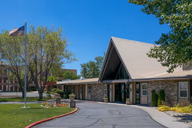Front exterior of a single-story rehabilitation facility with a peaked entrance, driveway, American flag, and landscaped lawn under a clear blue sky.