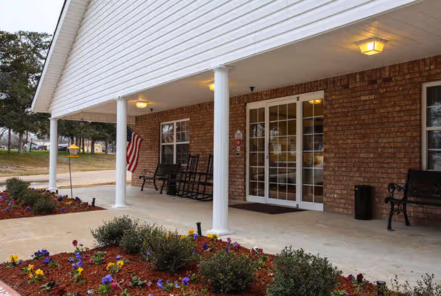 Covered entrance area of a building with white pillars, brick walls, and glass double doors. There are benches and rocking chairs along the wall, an American flag hanging near the entrance, and a flower bed with colorful flowers in the foreground.