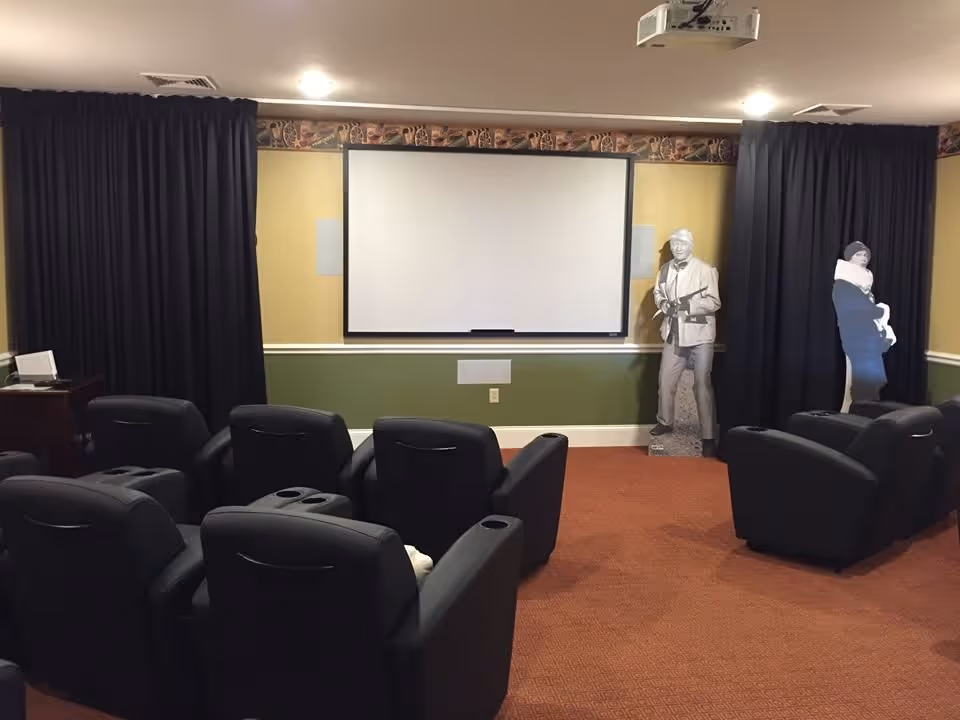 A small theater room with several black recliner chairs arranged in rows facing a large white projection screen on a yellow and green wall. There are black curtains on either side of the screen and two life-size cutouts of people standing near the right curtain. A projector is mounted on the ceiling.