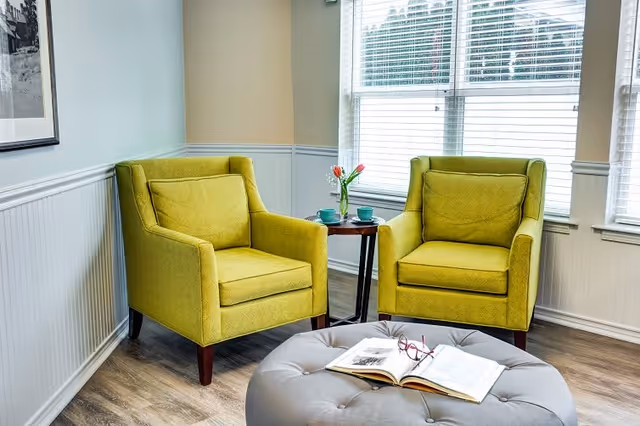 A cozy sitting area with two green upholstered armchairs facing each other, separated by a small round wooden table holding a vase with pink tulips and two teal cups. In front of the chairs is a large gray tufted ottoman with an open book and a pair of red reading glasses resting on it. The room has light-colored walls, white wainscoting, and large windows with white blinds letting in natural light.