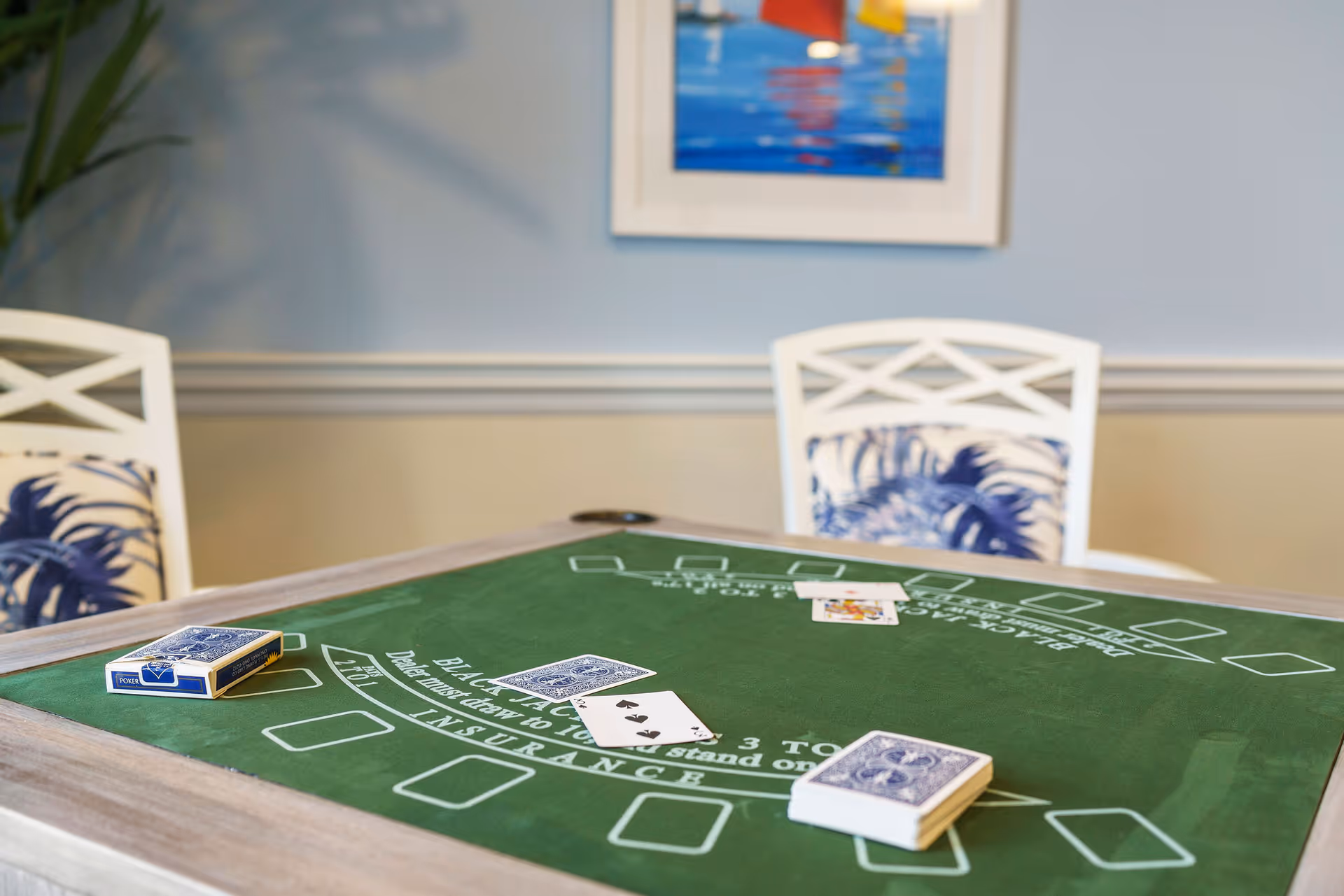 A green blackjack card table with playing cards and decks of cards on it, set in a room with light blue walls, white chairs with blue patterned cushions, and a framed colorful painting on the wall.