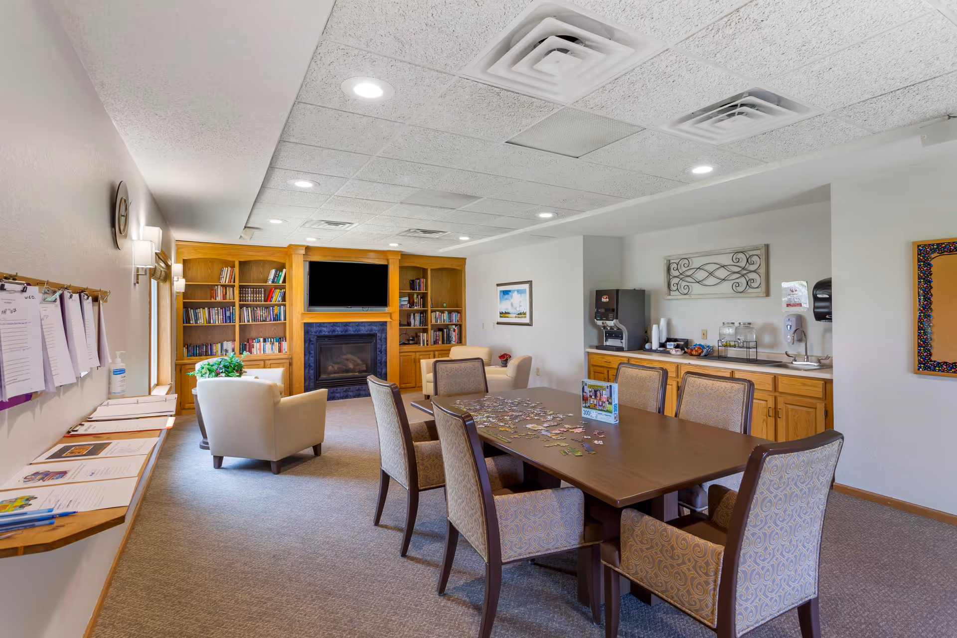 A cozy senior living common room with a large wooden table surrounded by six upholstered chairs. On the table, there is a partially completed jigsaw puzzle and a small display card. In the background, there is a built-in wooden bookshelf with books and a fireplace beneath a mounted flat-screen TV. To the right, there is a counter with a coffee machine, cups, and other refreshments. The room has carpeted floors, recessed ceiling lights, and a bulletin board on the wall.