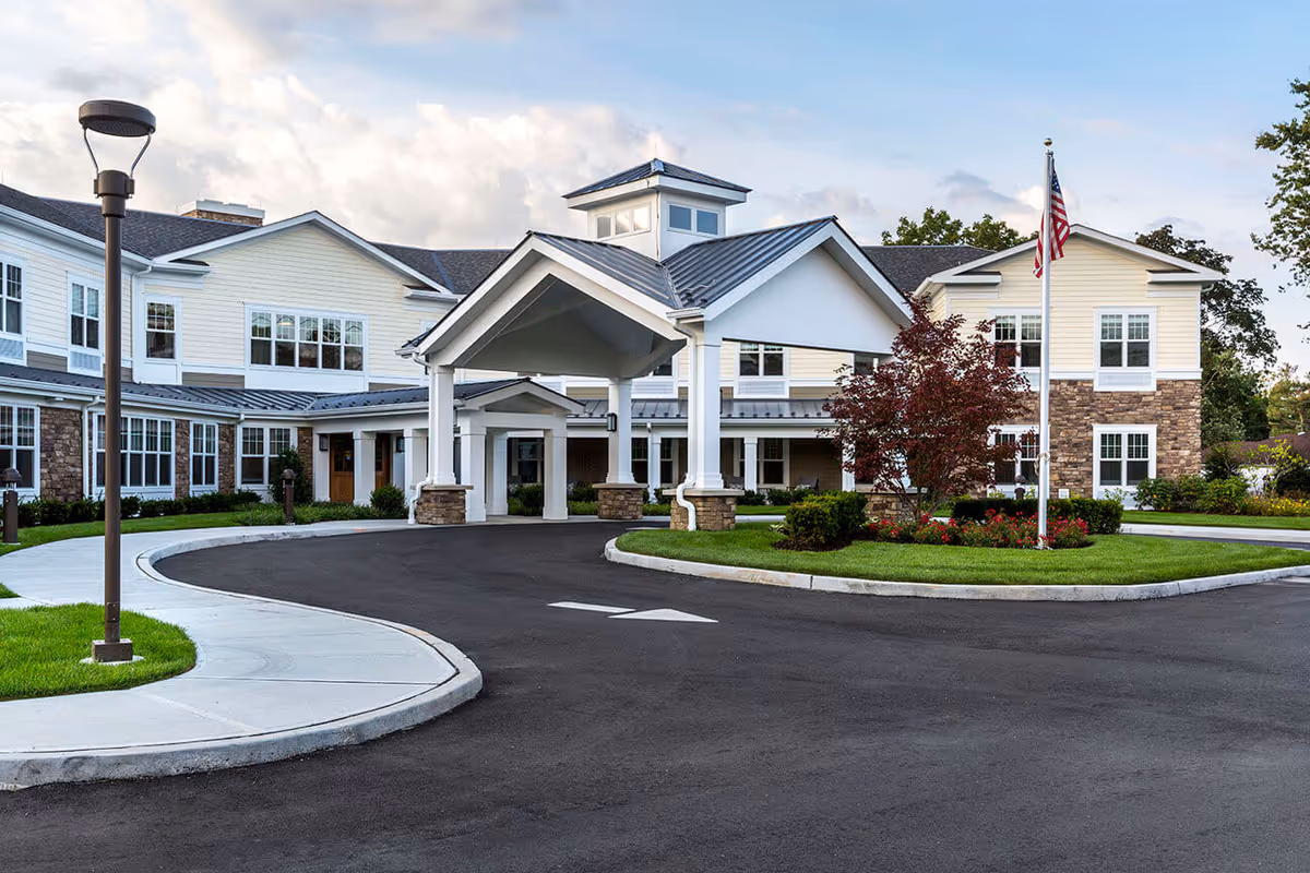 Exterior view of Orchard Estate of Woodbury, a senior living facility with a large covered entrance, landscaped greenery, and an American flag on a flagpole. The building features light-colored siding with stone accents and multiple windows.
