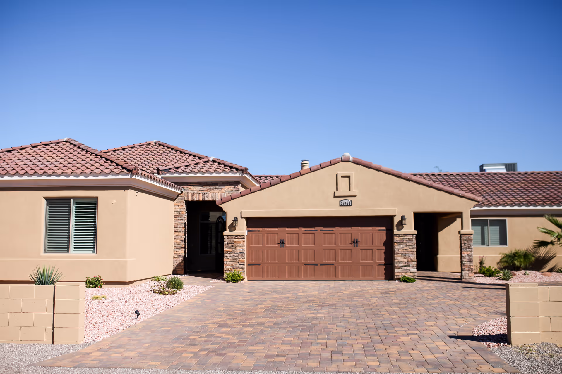 Front exterior view of a single-story house with a tiled roof, beige stucco walls, a double garage door, and a paved driveway under a clear blue sky.