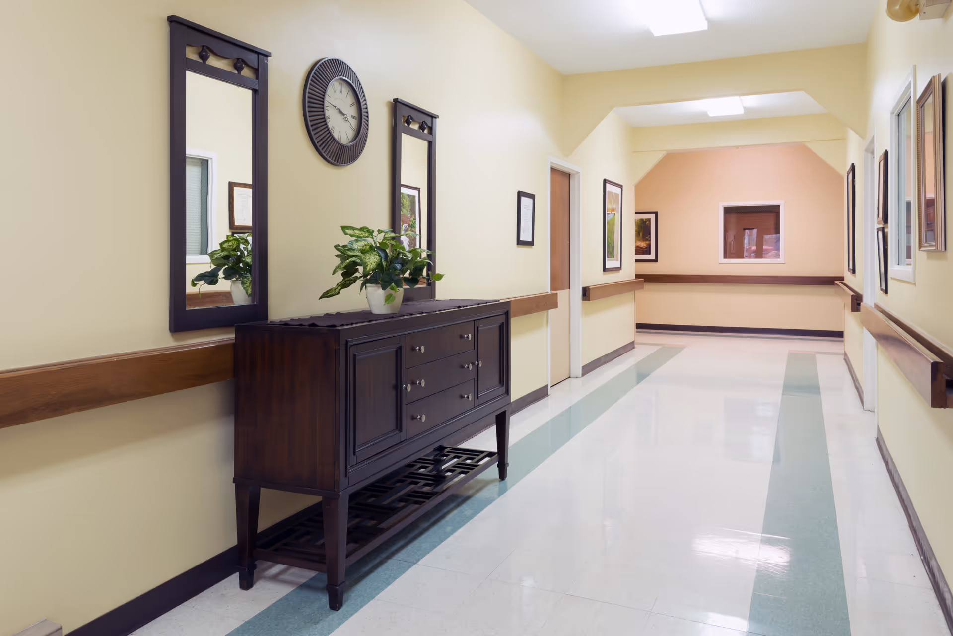 A clean, well-lit hallway in a care facility with light yellow walls and a shiny floor featuring green stripes. On the left side, there is a dark wooden console table with a potted plant on top, flanked by two vertical mirrors. A round wall clock is mounted between the mirrors. The hallway has handrails on both sides and framed pictures on the walls.
