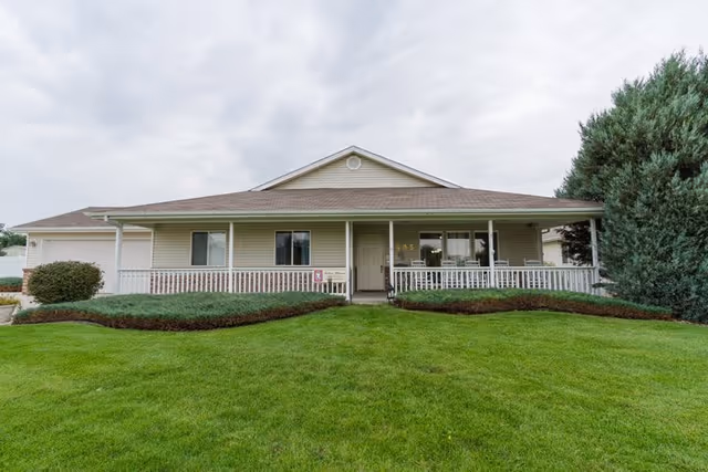 Single-story beige ranch-style building with a covered front porch, white railing, and a manicured green lawn.