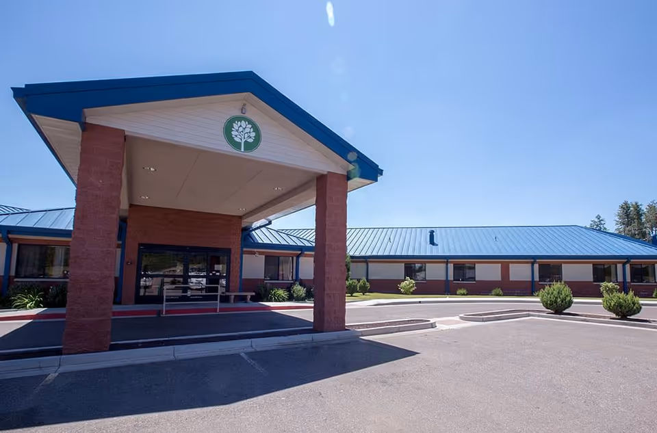 Exterior front view of Haven Health Lakeside facility with a covered entrance supported by two brick columns, blue metal roof, and a circular driveway with small bushes and clear blue sky.