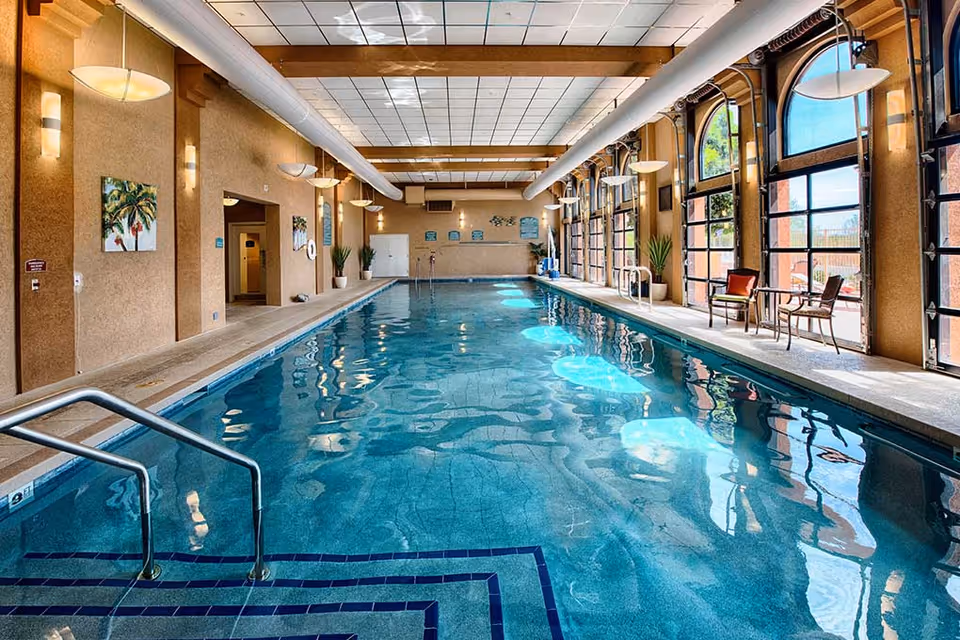 Indoor swimming pool with clear blue water, metal handrails, and steps leading into the pool. The pool area has large arched windows on the right side letting in natural light, with chairs and small tables placed along the windows. The walls are beige with decorative paintings and potted plants. The ceiling has white tiles with exposed beams and hanging light fixtures.