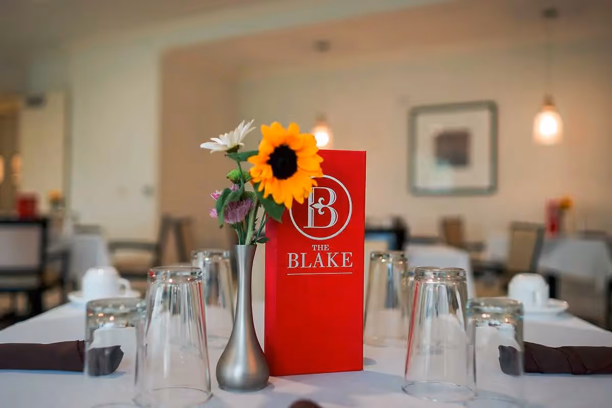 A dining table set with upside-down drinking glasses, white cups and saucers, and dark napkins. In the center of the table is a small silver vase with a sunflower and other flowers, next to a red menu card with the logo and text 'The Blake'. The background shows a softly lit dining room with tables and chairs.