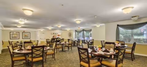 A bright senior living dining room with multiple round tables and place settings.