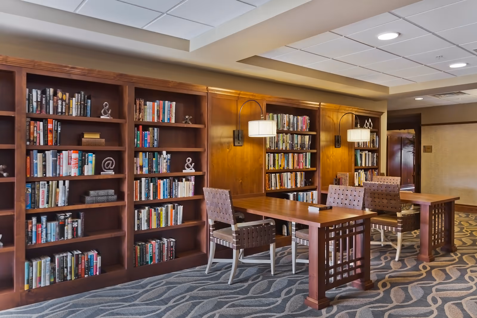 A cozy library or reading area with wooden bookshelves filled with books, two wooden tables, and four cushioned chairs. The room has patterned carpet flooring and wall-mounted lamps providing warm lighting.