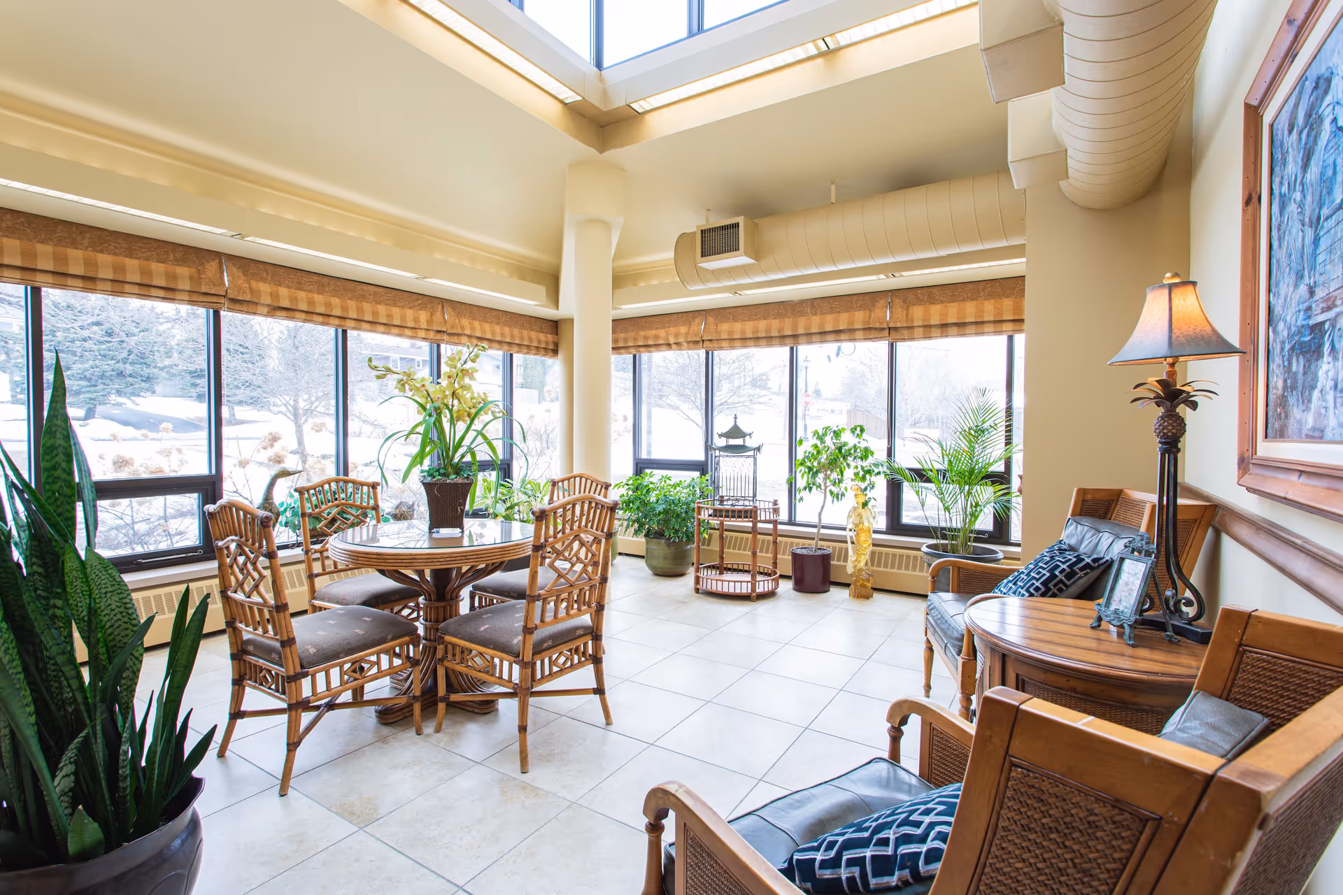A bright and airy common area with large windows letting in natural light. The room features a round wooden table with four cushioned chairs, several potted plants, two wooden armchairs with patterned cushions, a side table with a lamp, and a framed painting on the wall. The floor is tiled, and the ceiling has exposed ductwork.