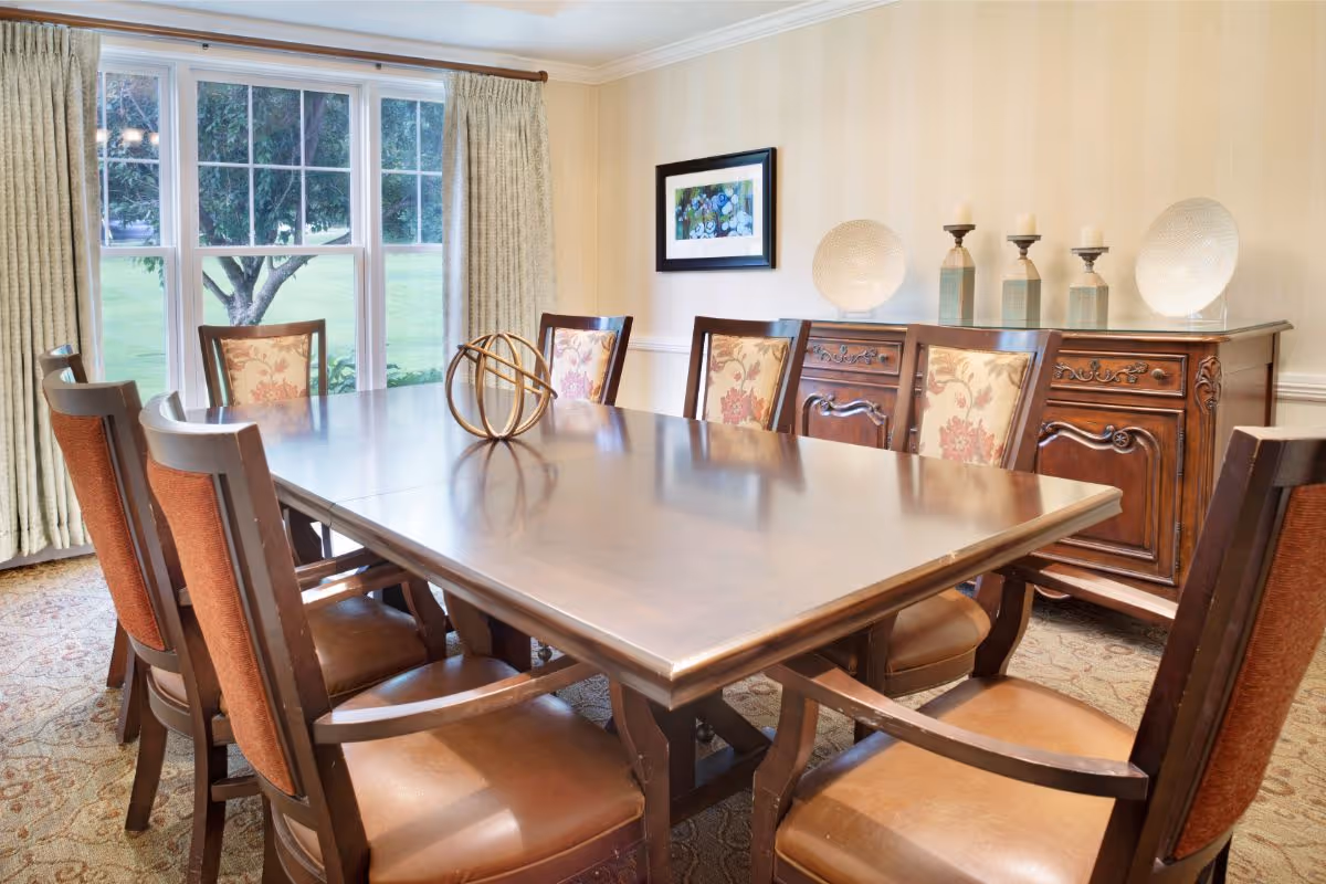 A dining room with a large wooden table surrounded by eight chairs, some with floral upholstery. Behind the table is a wooden sideboard with decorative plates and candles on top. A framed picture hangs on the wall above the sideboard. Large windows with beige curtains allow natural light to fill the room and show a green outdoor area with trees.