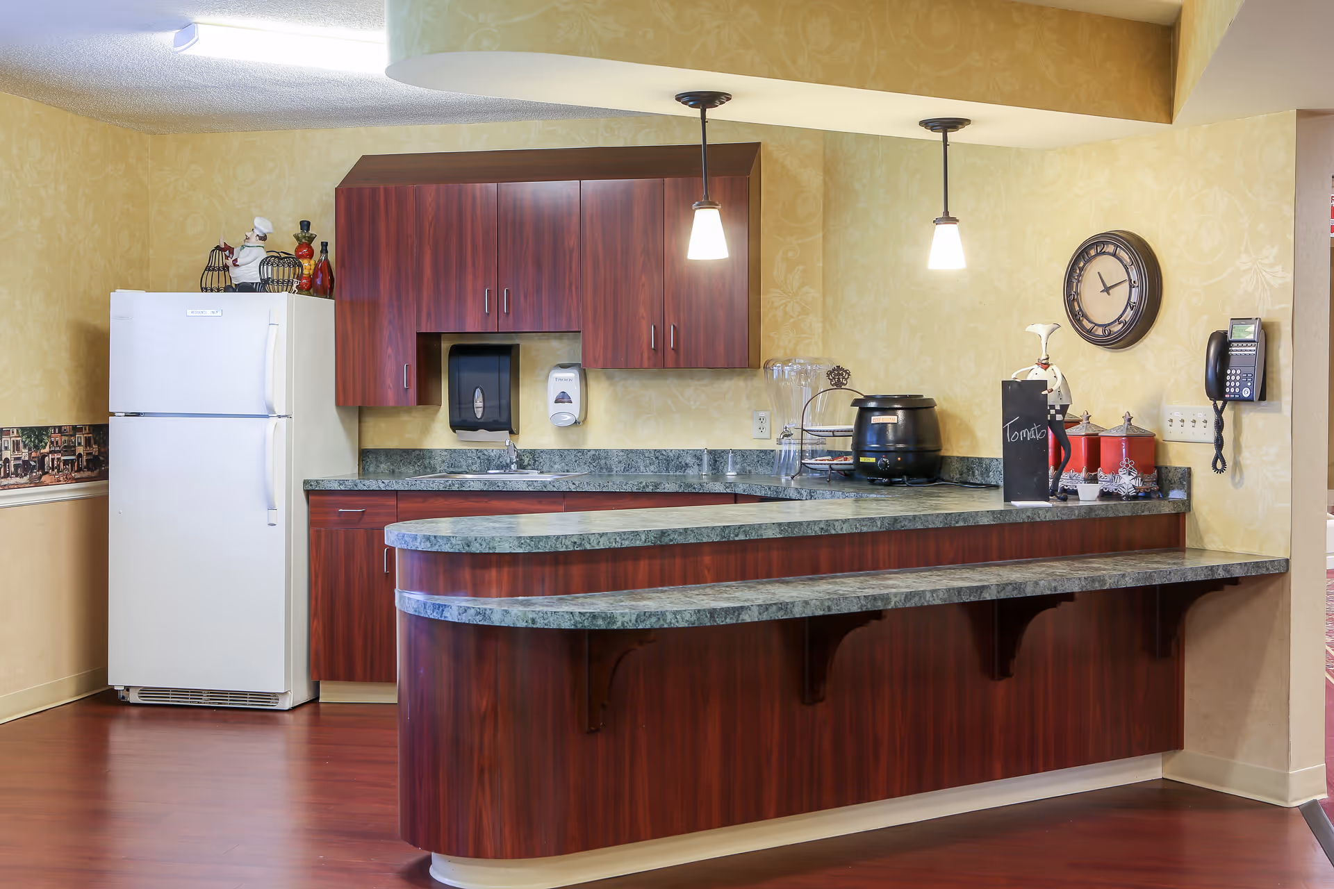 Interior view of a kitchen area in a senior living facility featuring a white refrigerator, dark wood cabinets, a green marble countertop with a curved breakfast bar, two hanging pendant lights, a wall clock, a telephone mounted on the wall, and various kitchen items including a soup pot and a chalkboard sign labeled 'Tomato'.