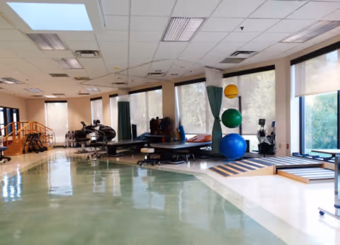 A spacious physical therapy or rehabilitation room with large windows letting in natural light. The room contains exercise equipment including therapy tables, large colorful exercise balls, parallel bars, and a small wooden staircase for mobility training. The floor is a mix of green and beige tiles, and the ceiling has fluorescent lighting.