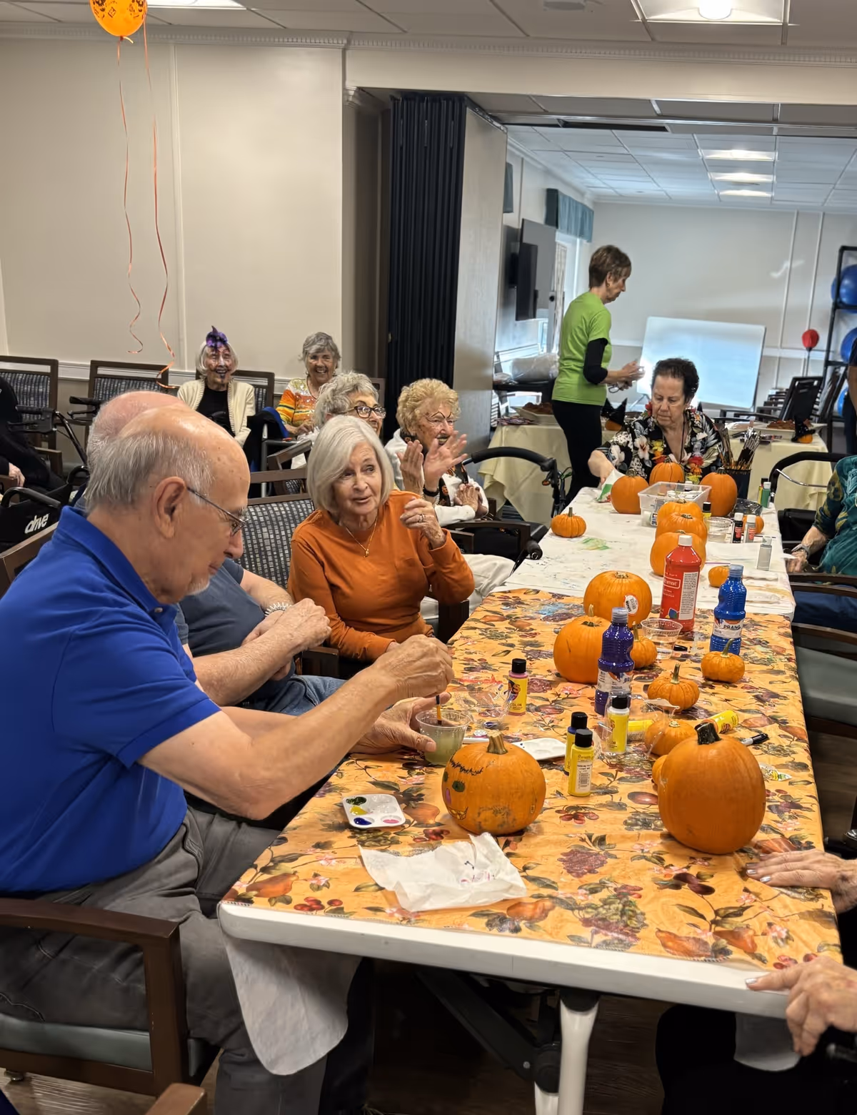 Seniors seated around a long decorated table painting small pumpkins in a communal activity room.