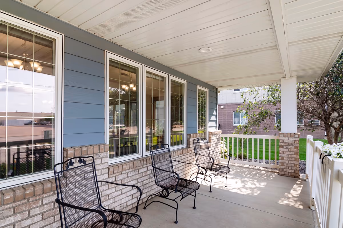 Covered outdoor patio area with three black metal chairs lined up against a brick and blue siding wall with windows. The patio has a white ceiling and railing, with greenery and a tree visible in the background.