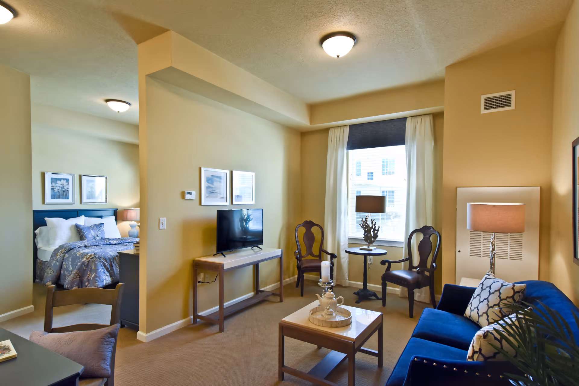 Open-plan living area with a blue sofa, coffee table, TV on a console, two chairs by a window, and a partially visible bedroom.