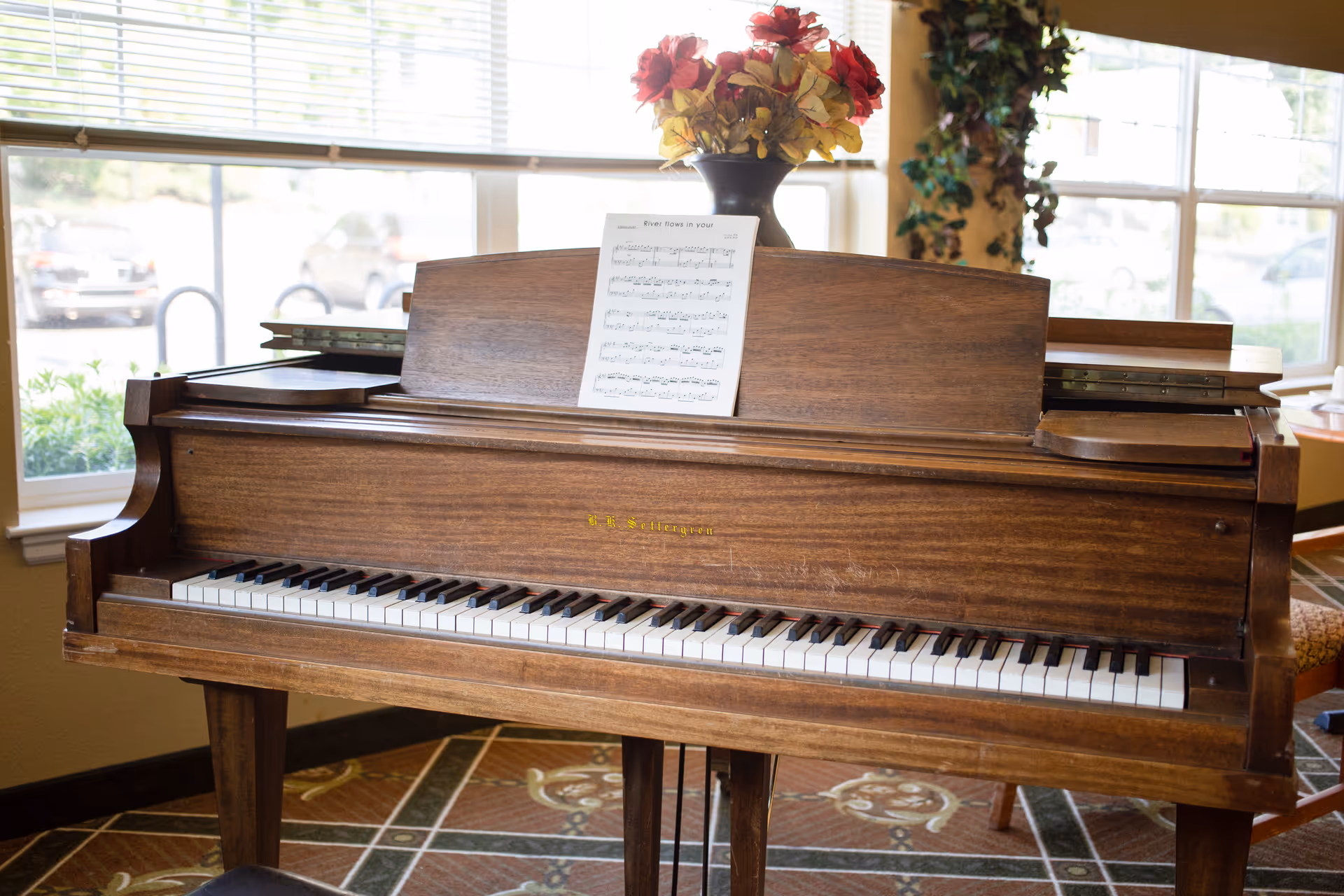 A wooden piano with sheet music placed on the music stand and a vase of red and yellow flowers on top. The piano is positioned in front of large windows with blinds, allowing natural light to fill the room. The floor has a patterned carpet, and some chairs and plants are visible in the background.