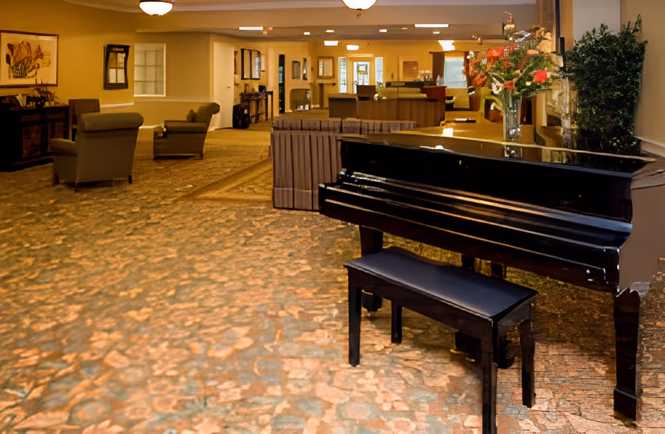 Interior view of a senior living facility lounge area featuring a black grand piano with a matching bench in the foreground. The room has carpeted floors, several armchairs, a table with a flower vase, and warm lighting fixtures on the ceiling. The space appears cozy and inviting with framed artwork on the walls and a mix of seating arrangements.