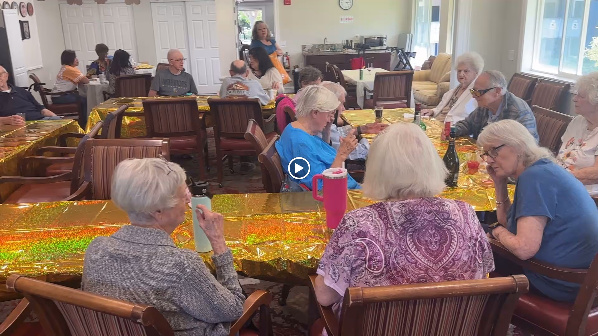 Seniors seated around tables covered with gold tablecloths in a communal dining/activity room.