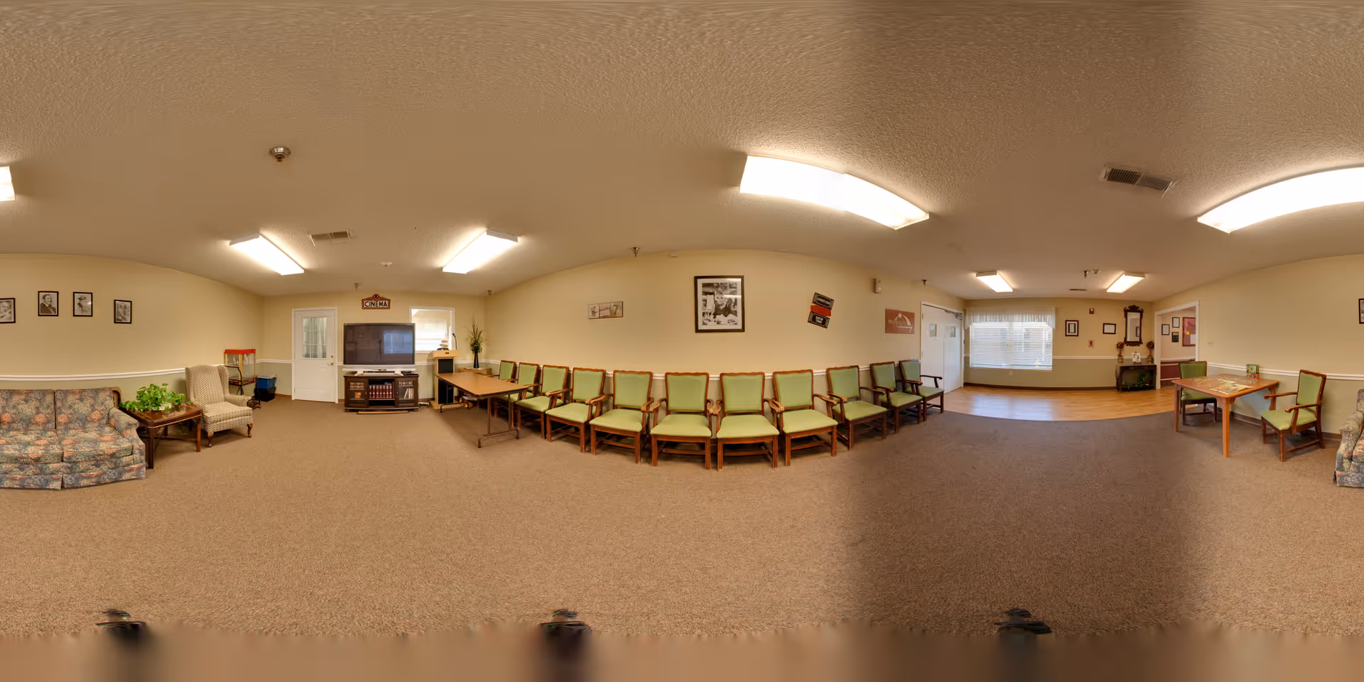A spacious common area in a senior living facility with beige walls and carpeted floors. The room features a row of green cushioned wooden chairs lined up against the wall, a floral patterned sofa, an armchair, a small table with a plant, a television on a stand, and a few framed pictures on the walls. The room is well-lit with ceiling lights and has a door and windows letting in natural light.