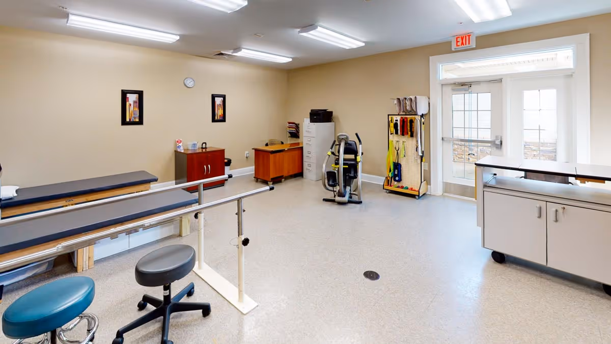 A physical therapy or rehabilitation room with parallel bars for walking exercises, two padded benches, two stools, a desk with a chair, a filing cabinet, exercise equipment, and a rack holding various therapy tools. The room has beige walls, a clock, two framed pictures, and a glass door with an exit sign above it.
