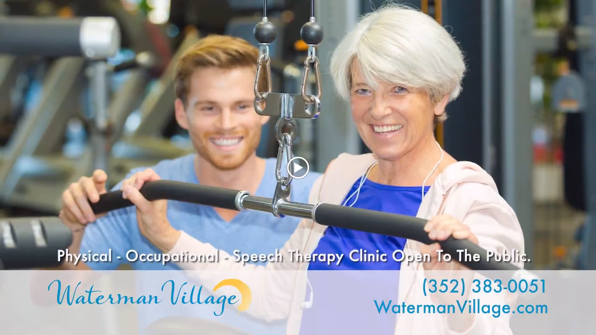 An elderly woman with white hair is smiling while using a lat pulldown machine in a gym, assisted by a younger male trainer who is also smiling. The background shows gym equipment.