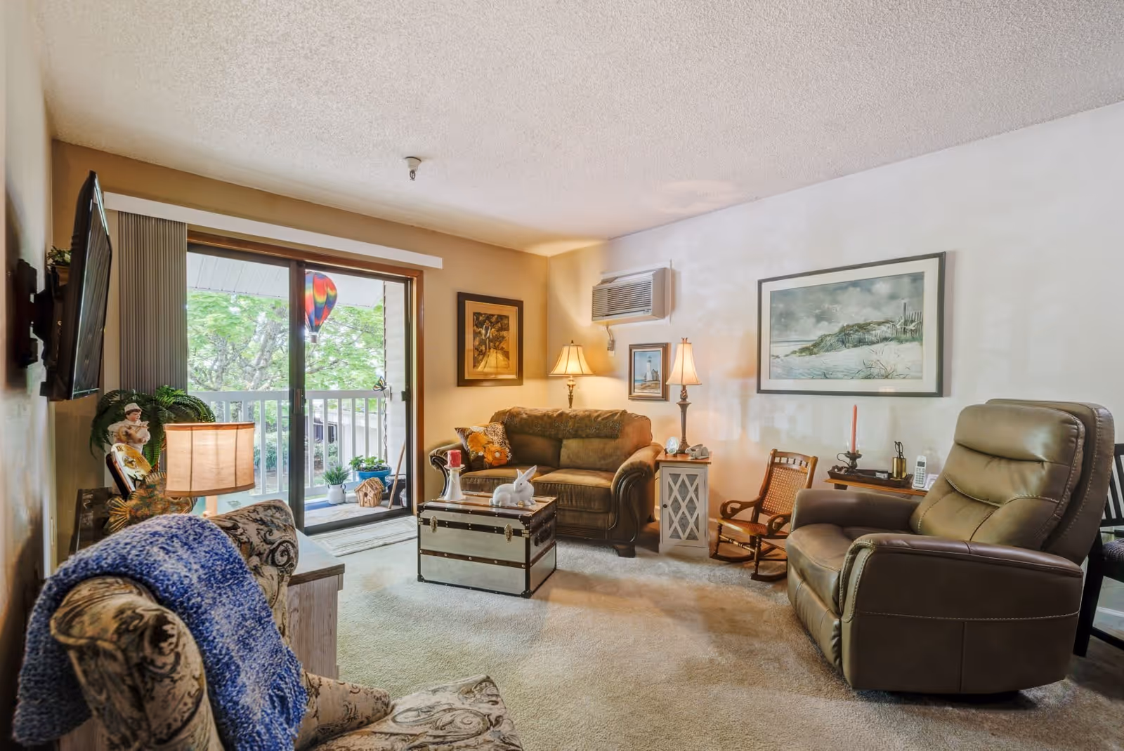 A cozy living room with a brown leather recliner, a patterned armchair with a blue throw, a brown sofa, and a trunk-style coffee table. The room has two table lamps, framed artwork on the walls, and a sliding glass door leading to a balcony with plants and a colorful hot air balloon decoration visible outside.