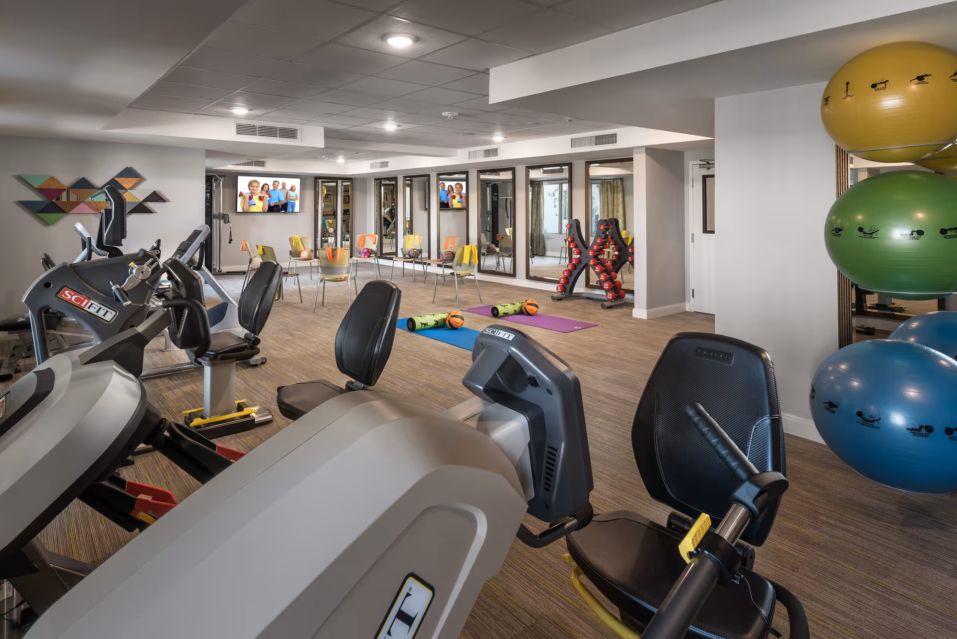 A fitness room in a senior living facility featuring exercise equipment such as stationary bikes and weight machines. The room has large mirrors on one wall, colorful exercise balls stacked on a rack, yoga mats, foam rollers, and small basketballs on the floor. Several chairs are arranged in a semi-circle facing two wall-mounted TVs displaying images of smiling seniors. The room has a modern design with recessed lighting and wood-style flooring.