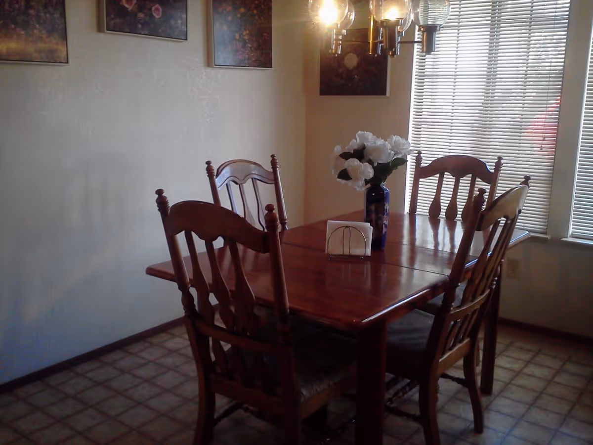 A wooden dining table with four chairs, a vase of white flowers and a napkin holder under a hanging light fixture beside windows with blinds.