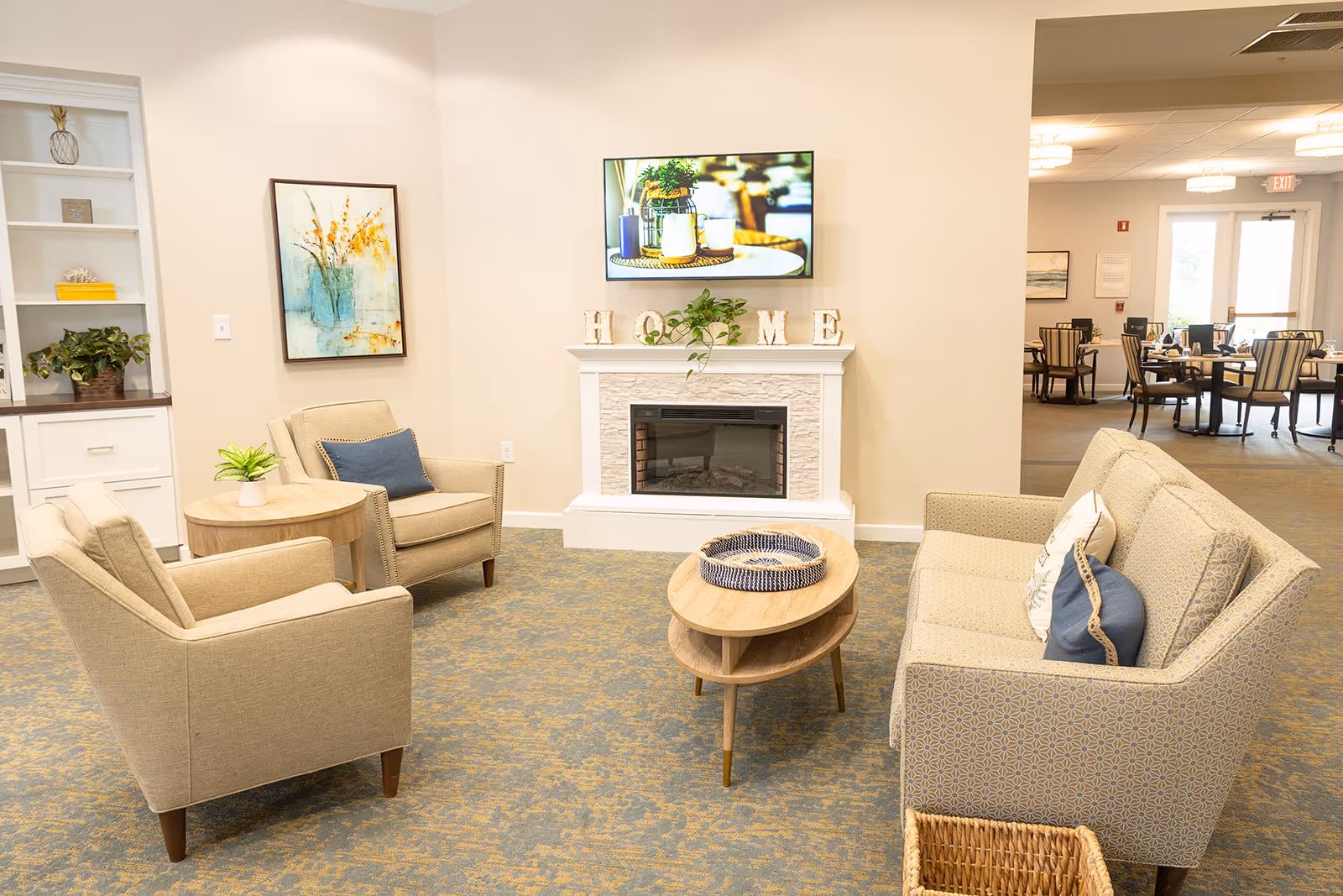 A cozy senior living facility common area with beige armchairs and a patterned sofa arranged around a wooden coffee table. A white fireplace with decorative letters spelling 'HOME' and a green plant sits beneath a wall-mounted TV displaying a decorative scene. In the background, a dining area with tables and chairs is visible through an open doorway.