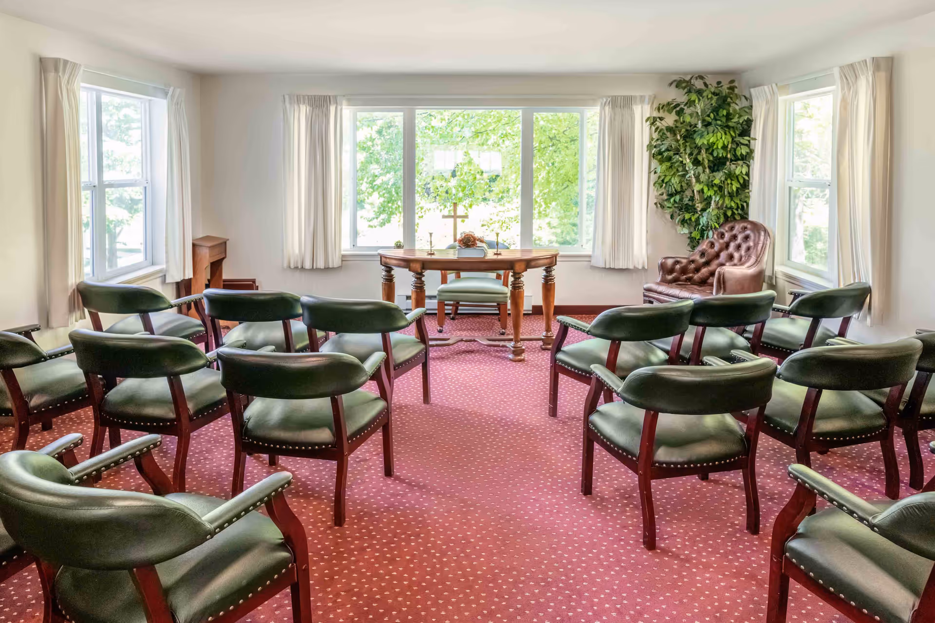 A small meeting or chapel room with green cushioned chairs arranged in rows facing a wooden table with a cross and decorative items on it. The room has large windows with white curtains letting in natural light, a red carpet with a dotted pattern, and a brown leather armchair next to a tall green plant in the corner.