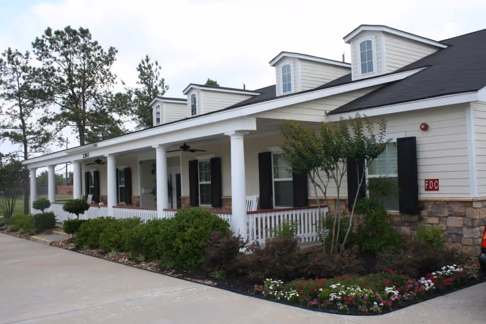 Exterior view of a single-story building with white siding, black shutters, and a covered porch supported by white columns. The porch has rocking chairs and ceiling fans. There are well-maintained shrubs and flowers along the front, with a concrete driveway or walkway beside it. Tall pine trees are visible in the background under a partly cloudy sky.