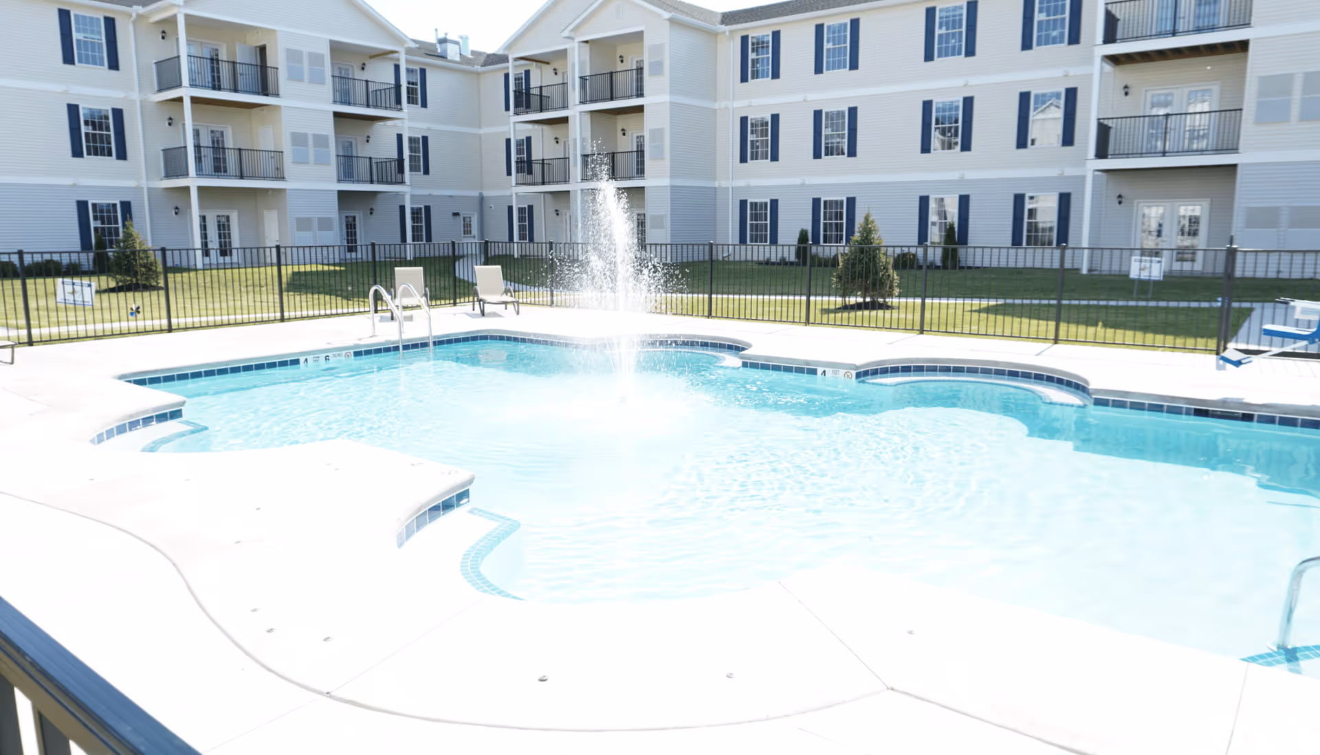 Outdoor swimming pool with a water fountain in the center, surrounded by a concrete deck and a black metal fence. In the background, there is a multi-story residential building with balconies and windows, and some lawn chairs placed near the pool.