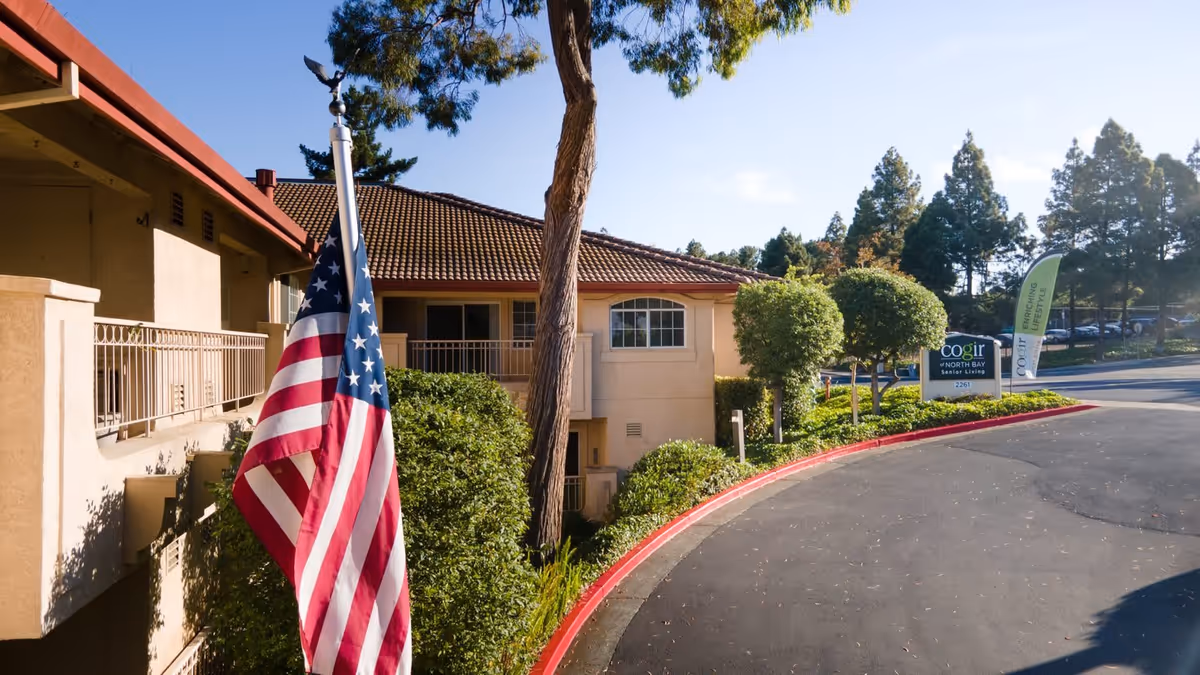 Exterior view of Cogir of North Bay Senior Living facility with an American flag in the foreground, trees, bushes, and a curved driveway leading to the building under a clear sky.