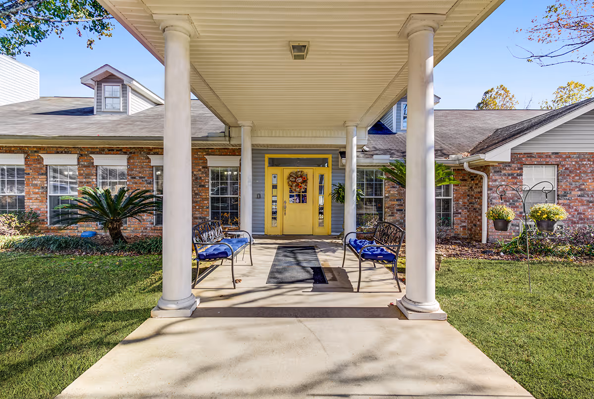 Front entrance of Marigold House featuring a covered porch with four white columns, two metal benches with blue cushions, a yellow door decorated with a wreath, brick exterior walls, and green lawn with plants and flowers.