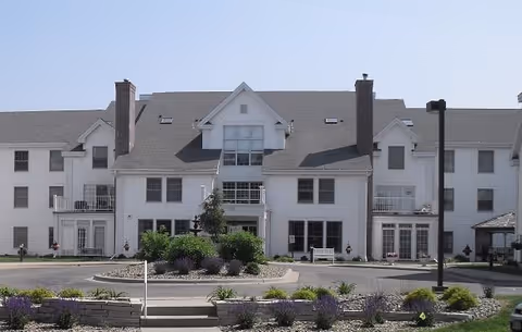 Front exterior view of a large white senior living facility building with multiple windows, balconies, and chimneys. There is a circular driveway with landscaped greenery and flowers in front of the building.