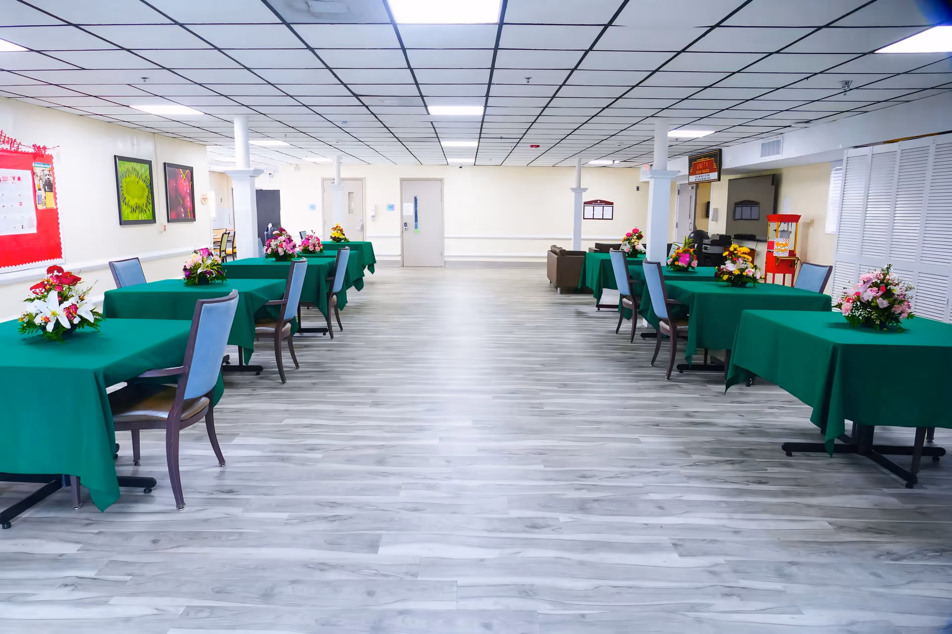 A spacious dining area with several tables covered in green tablecloths, each adorned with a floral centerpiece. Chairs are arranged around the tables, and the room has light-colored walls, a tiled ceiling, and wood-patterned flooring. There is a popcorn machine and some seating in the background.