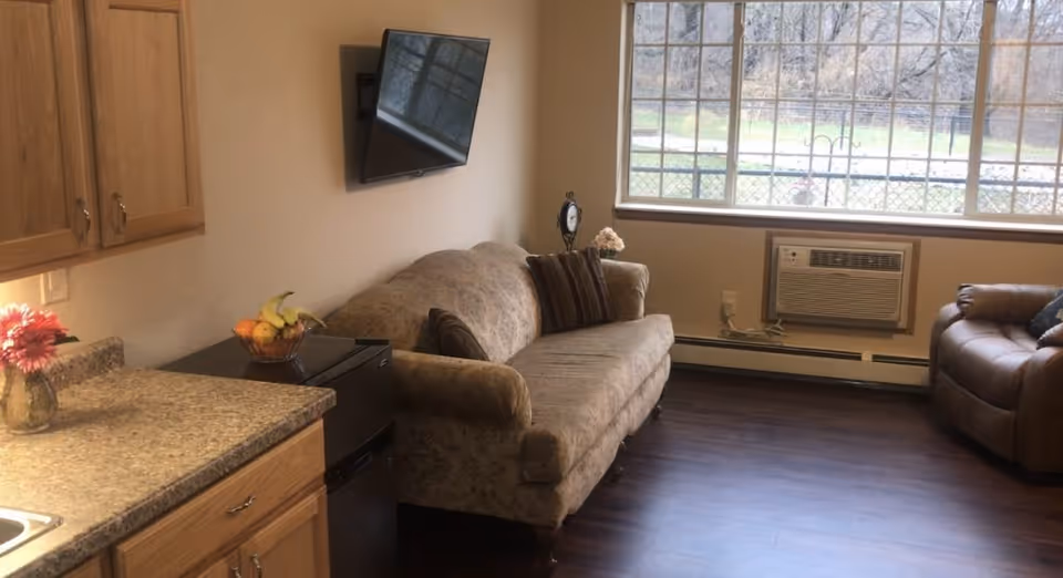 A cozy living area in SpringBrook Assisted Living featuring a floral-patterned sofa with two striped pillows, a brown leather armchair, a wall-mounted flat-screen TV, a large window with a view of the outdoors, an air conditioning unit below the window, and a kitchen counter with wooden cabinets, a small black refrigerator, a bowl of fruit, and a vase with a pink flower.