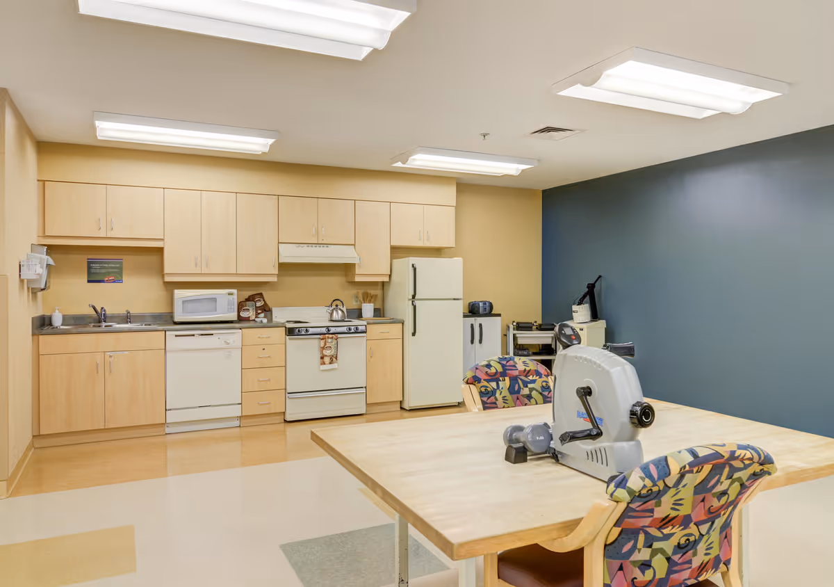 A small kitchen area with light wood cabinets, a white refrigerator, stove, microwave, dishwasher, and sink. In the foreground, there is a wooden table with two colorful patterned chairs and exercise equipment including hand weights and a pedal exerciser. The walls are painted beige and blue, and the room is lit by fluorescent ceiling lights.