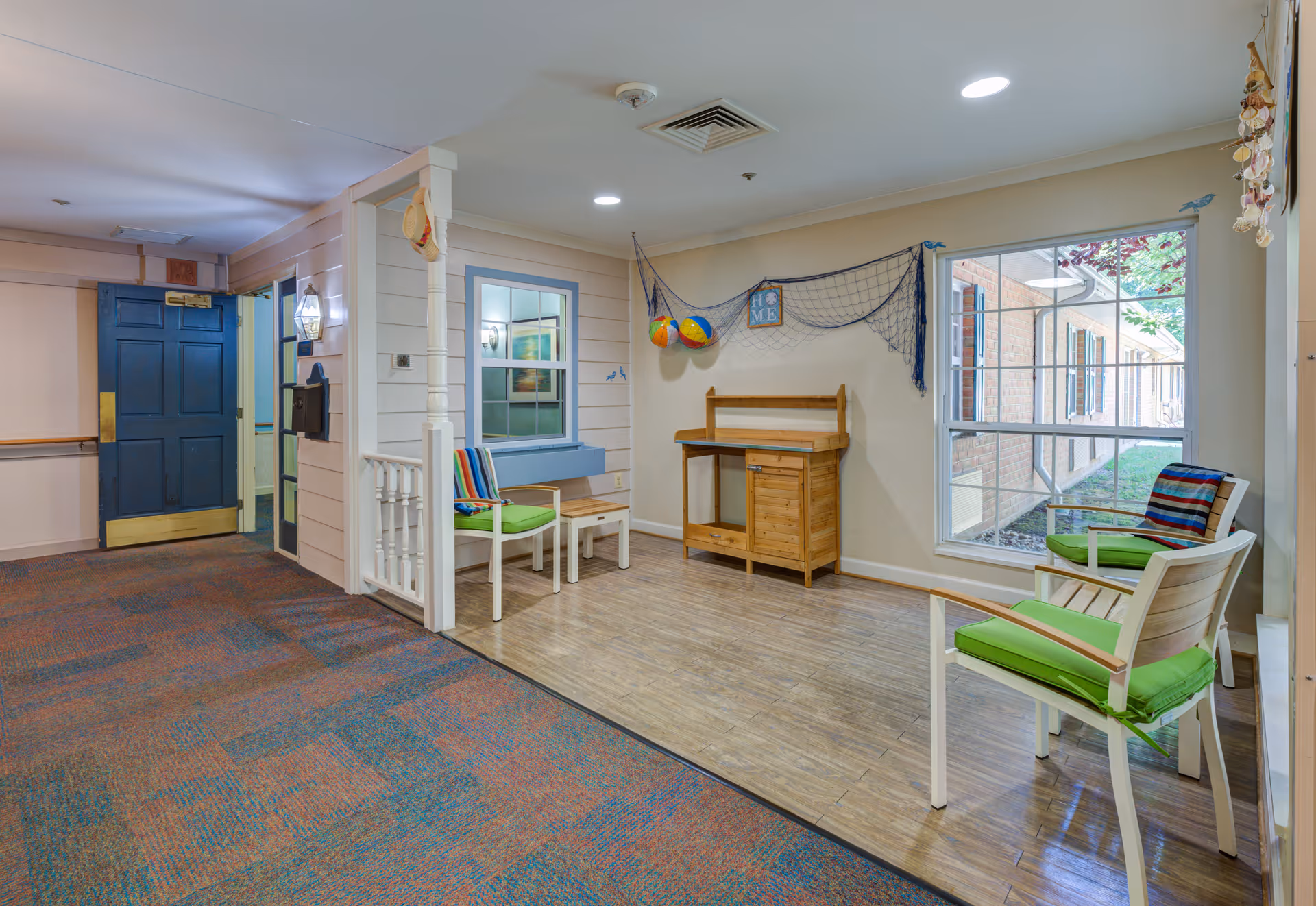 A cozy indoor seating area with two white chairs featuring green cushions and colorful striped pillows, a small wooden table, and a wooden cabinet against the wall. The wall is decorated with a fishing net and two hanging beach balls. A large window shows an exterior view of a brick building and greenery. The floor is a combination of carpet and wood, and a blue door is visible in the background.