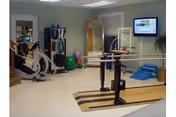 A rehabilitation room with exercise equipment including parallel bars, an elliptical machine, exercise balls, a mirror, and a wall-mounted television screen.