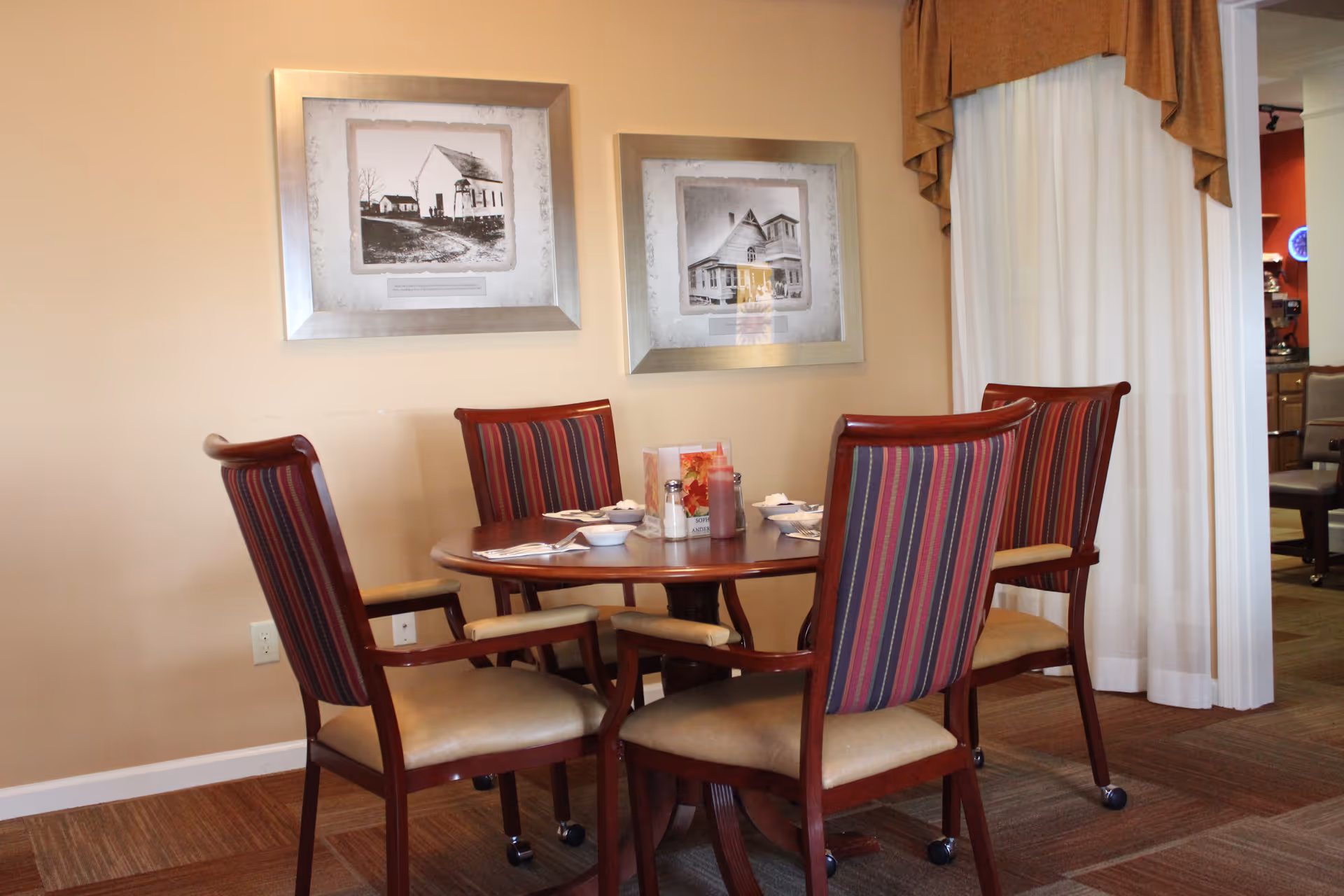 A round wooden dining table set with four chairs featuring striped upholstery and beige cushions. The table has place settings including bowls, napkins, and condiments. Two framed black and white photographs hang on the beige wall behind the table. A window with white curtains and a brown valance is visible to the right.