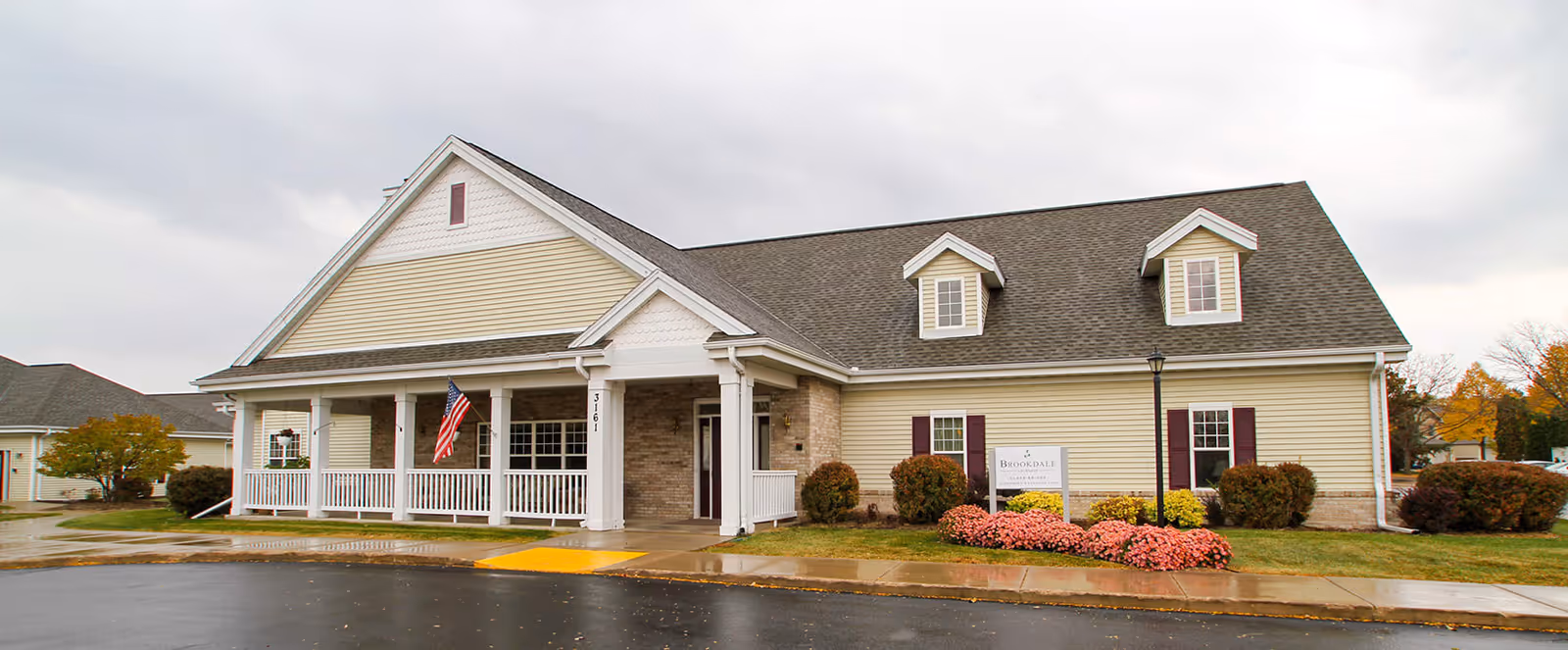 Exterior view of a single-story building with beige siding, white trim, and a gray shingled roof. The building has a covered porch with white railings and columns, an American flag hanging near the entrance, and landscaping with bushes and flowers in front. A sign near the entrance reads Brookdale LaCrosse.