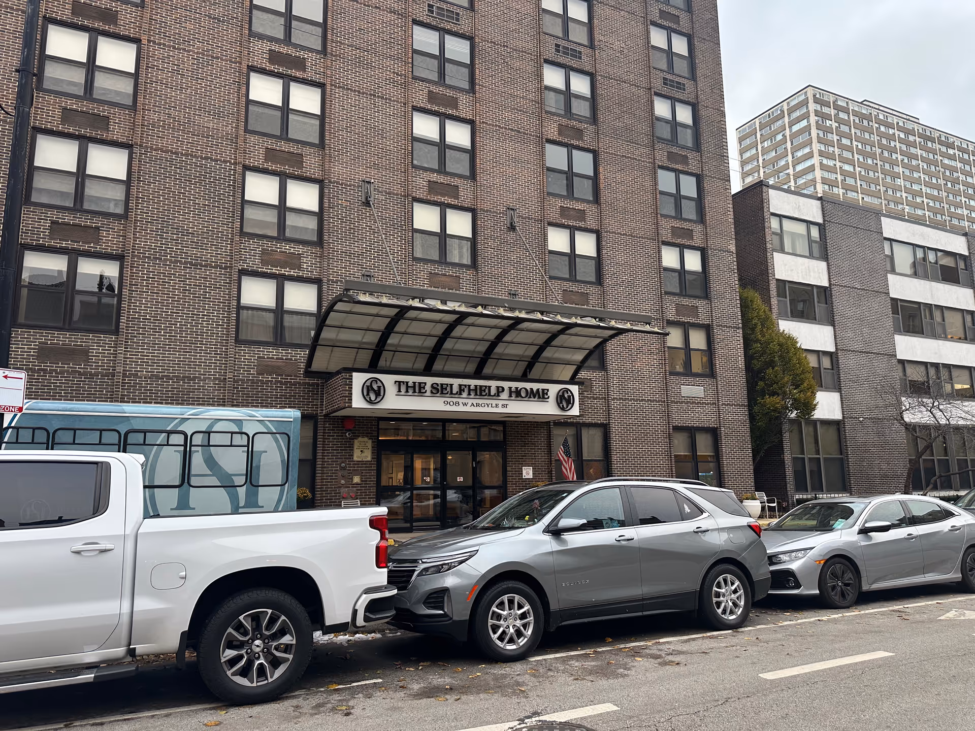 Exterior view of The Selfhelp Home Senior Living Community building with a brick facade, multiple windows, and a covered entrance. Several cars are parked along the street in front of the building.