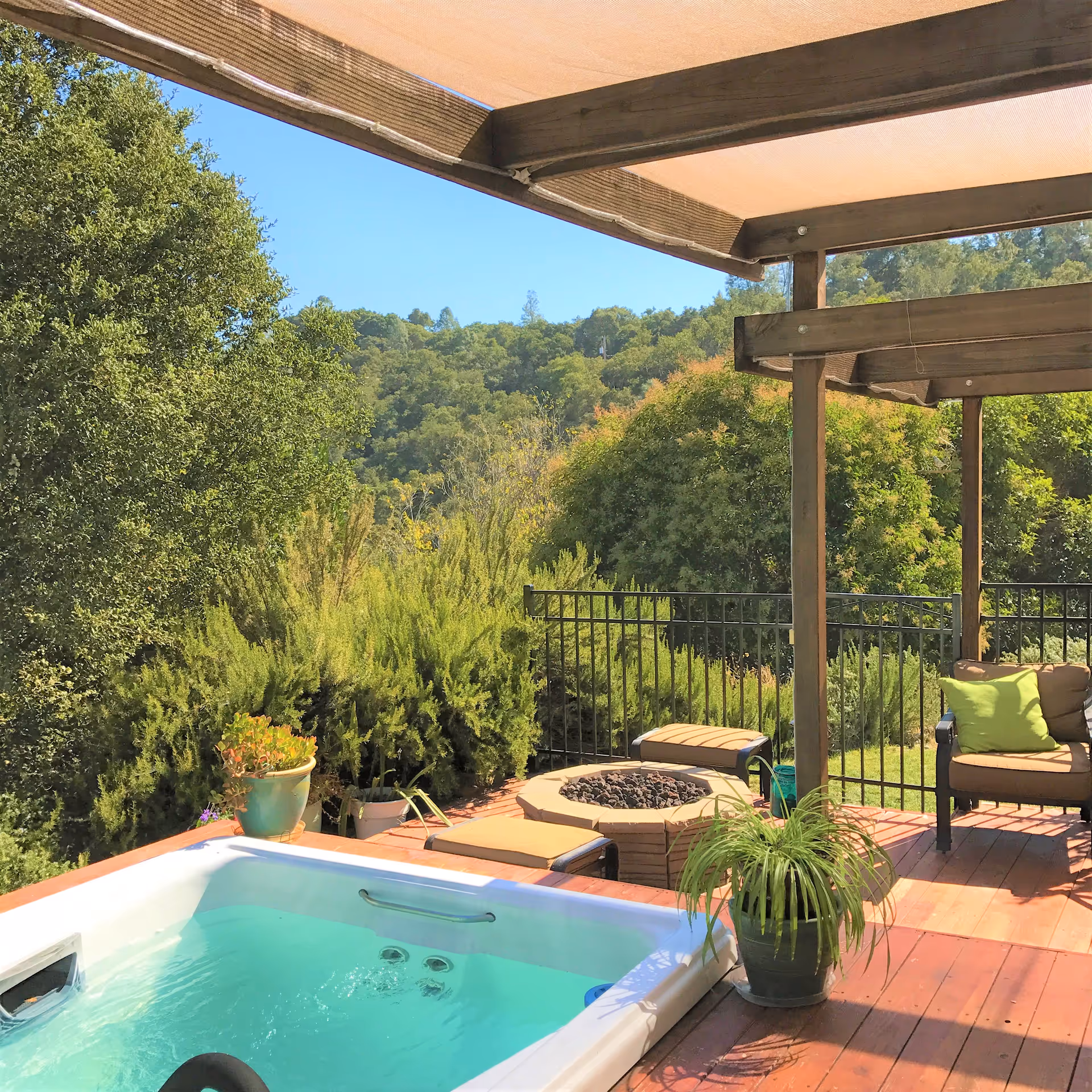 Outdoor patio area with a hot tub, cushioned seating, a fire pit, potted plants, and a wooden pergola overhead. The background features lush green trees and a clear blue sky.