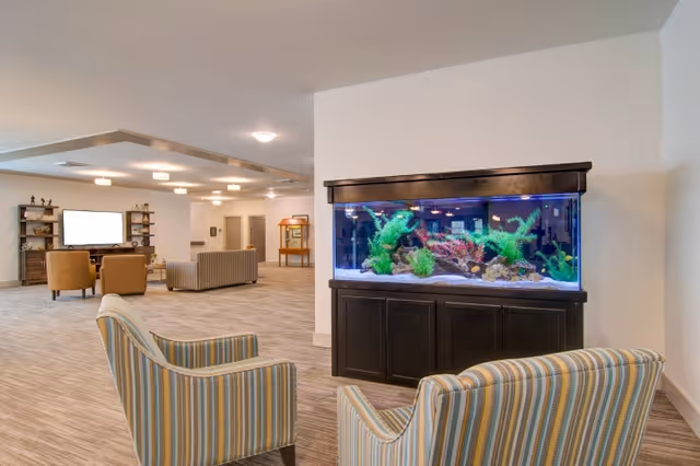A spacious living area in a senior living facility featuring a large aquarium with colorful aquatic plants and rocks, two striped armchairs in the foreground, and additional seating including a sofa and chairs arranged around a television in the background. The room has neutral-colored walls and carpeted flooring with ceiling lights providing illumination.