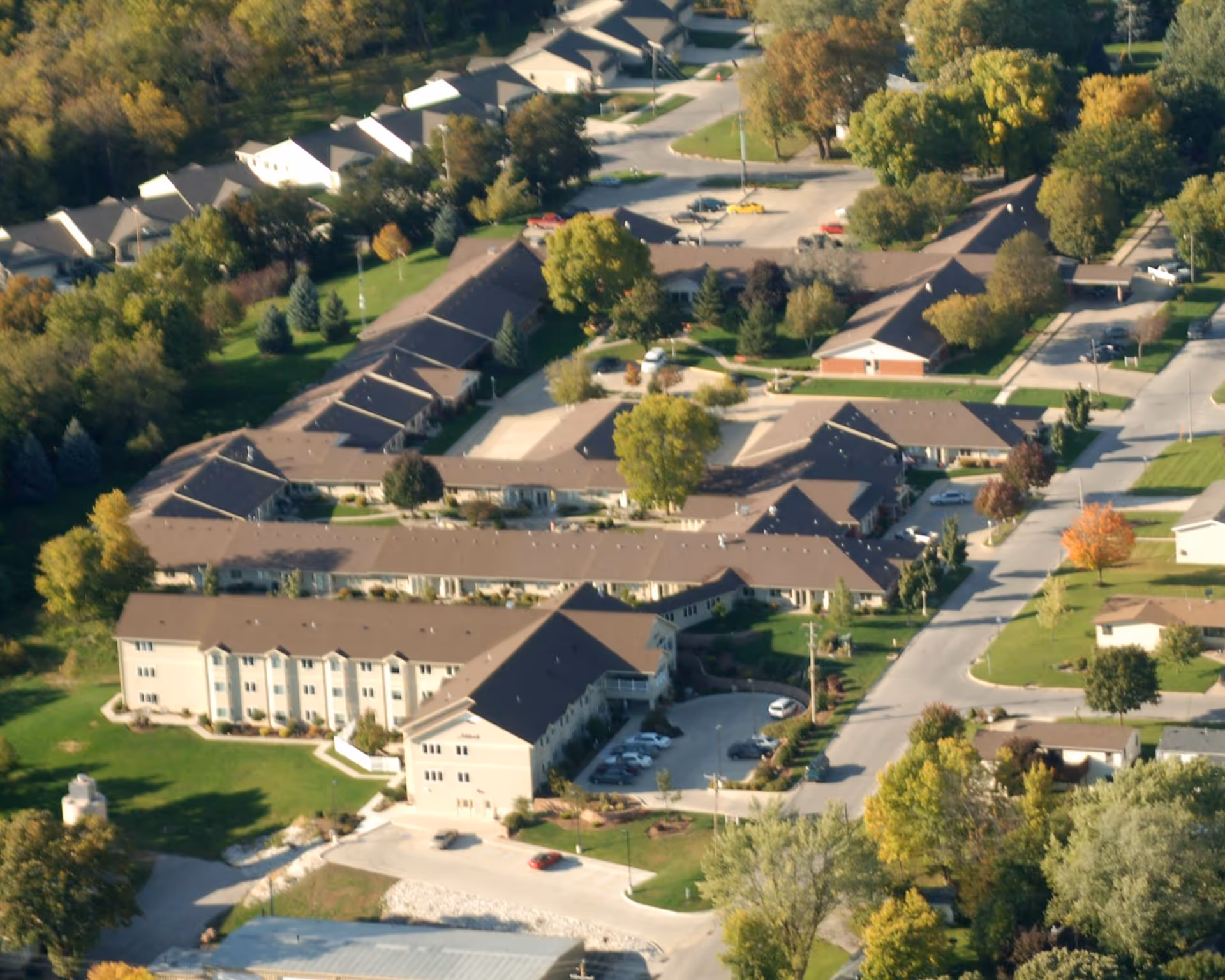 Aerial view of a senior living community with multiple connected buildings surrounded by trees and parking areas, showing a peaceful neighborhood setting with green lawns and roads.